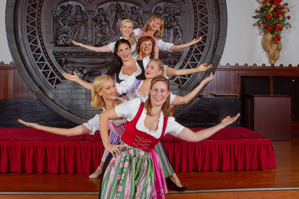 Austrian Dinner Show, Vienna, seven women and colorful dirndls stand in a row and look alternately left and right with outstretched arms into the camera