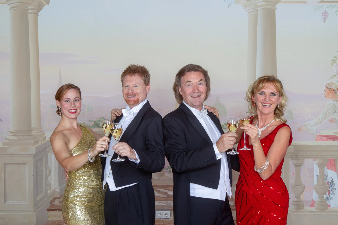 Austrian Dinner Show, Vienna, two women in gold and red evening dresses standing next to two men in black tails, clinking champagne glasses and smiling at the camera