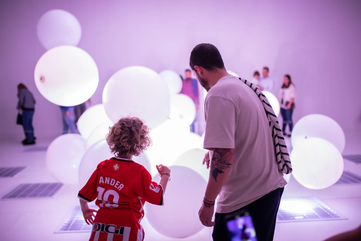 A man and a child marvel at large white balloons flying around. Other visitors can be seen in the background.