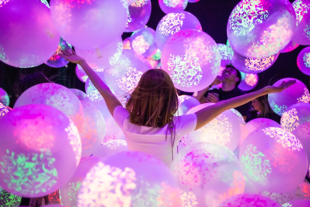 A girl stands with her back to the camera between glowing pink balloons and raises her arms in the air.