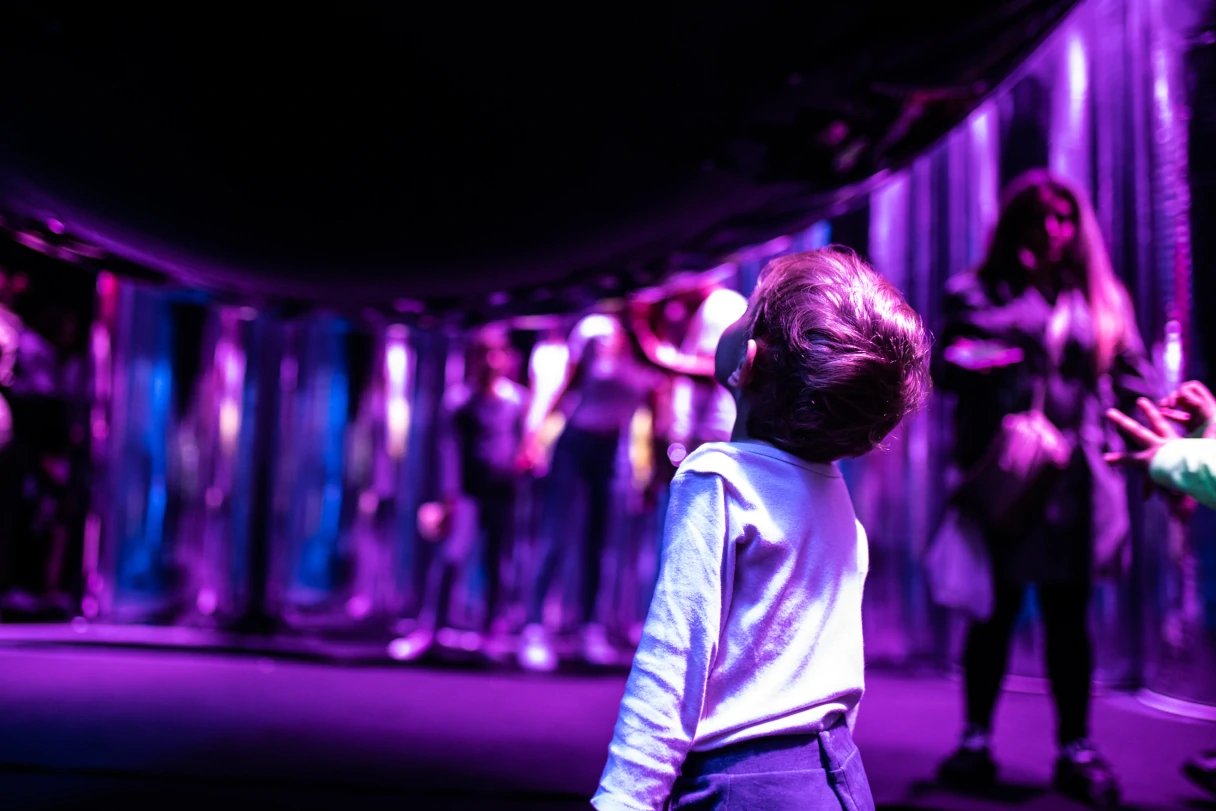 Visitors to the Balloon Museum look around the room. Pink light and a small child stands with its back to the camera and looks up at the ceiling.