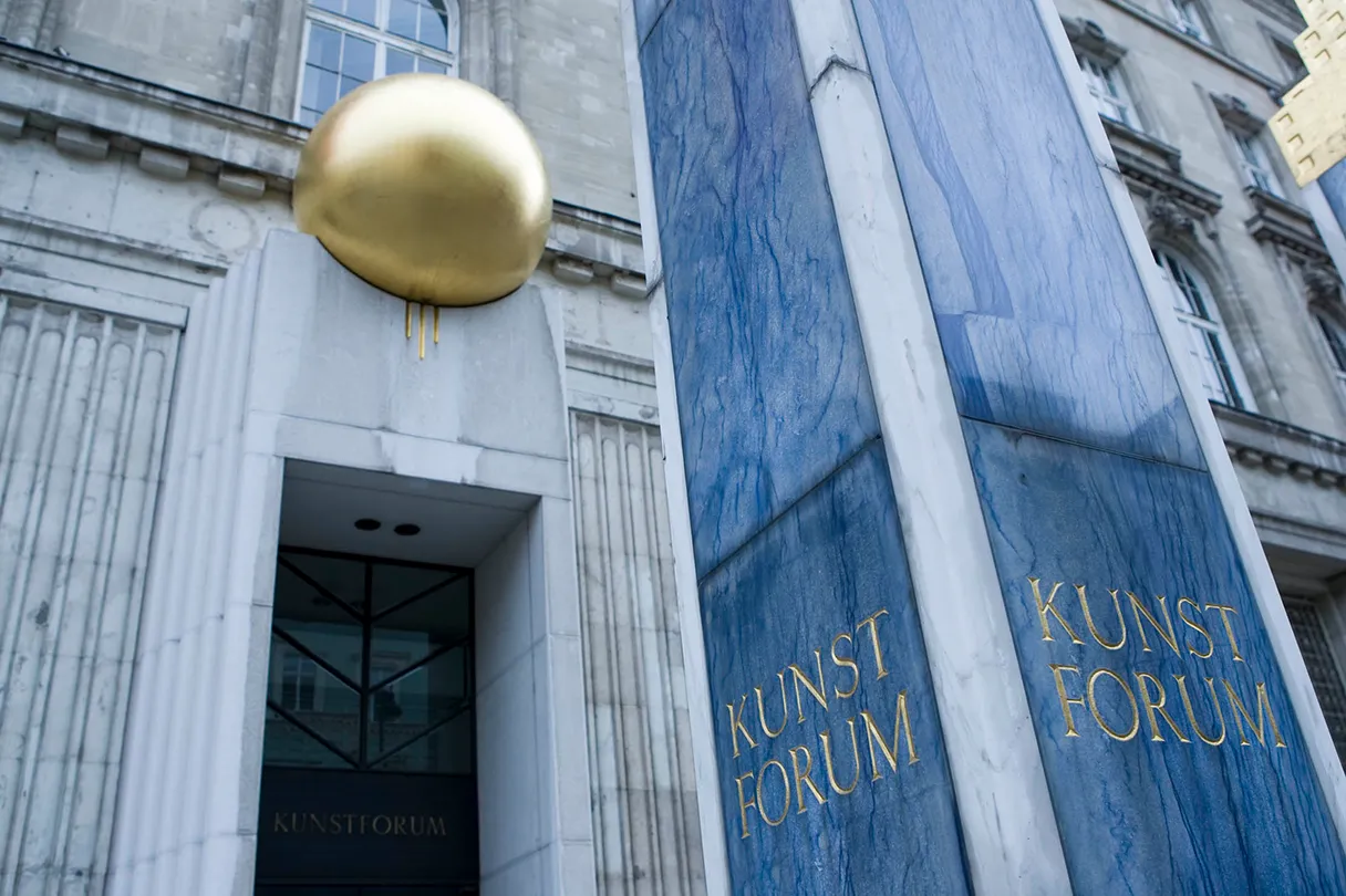 Exterior facade of the Kunstforum Wien. A blue marble column on which Kunstforum is written in gold lettering can be seen on the right edge of the picture. A golden sphere above a brown door built into the façade of the building.