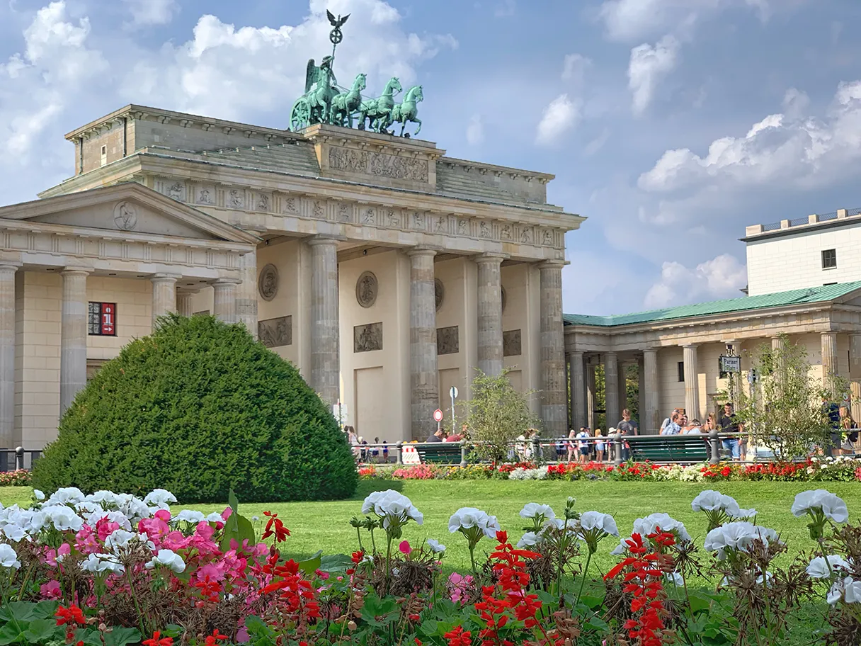 Original Berlin walks, Blick auf das Brandenburger Tor, im Vordergrund sind Blumenrabatten mit weiß-roten Blumen, grüner Busch wächst auf der grünen Wiese