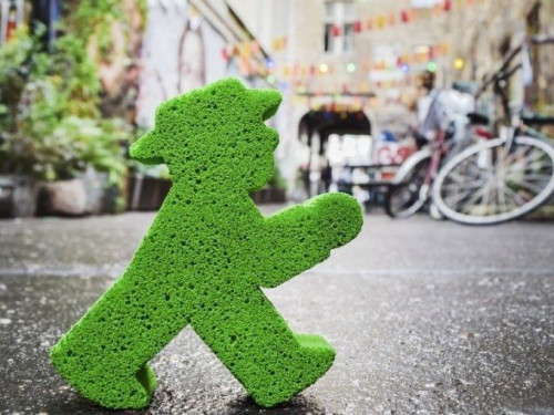 Green sponge in the shape of the Ampelmännchen figure in the foreground, on a wet street near Hackesche Höfe, Berlin; bicycles and colorful lights in the background.