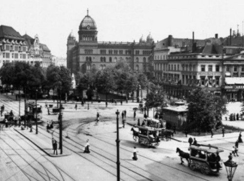 Black and white photo of Berlin's Alexanderplatz around 1908 with horse-drawn carriages, trams, a large building in the background and people crossing the street.
