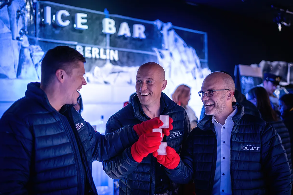 Berlin Icebar, drei Männer stoßen in der Icebar mit ihren Eis-Gläsern an, im Hintergrund ist die Eisbar zu erkennen