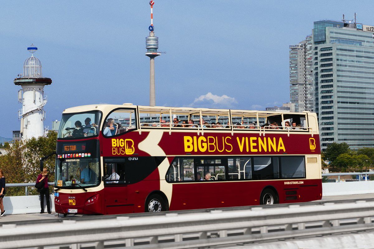 Big Bus Tours Vienna, bordeauxfarbener Sightseeingbus fährt auf der Straße, im Hintergrund sind der Leuchtturm und Donauturm Wien zu sehen