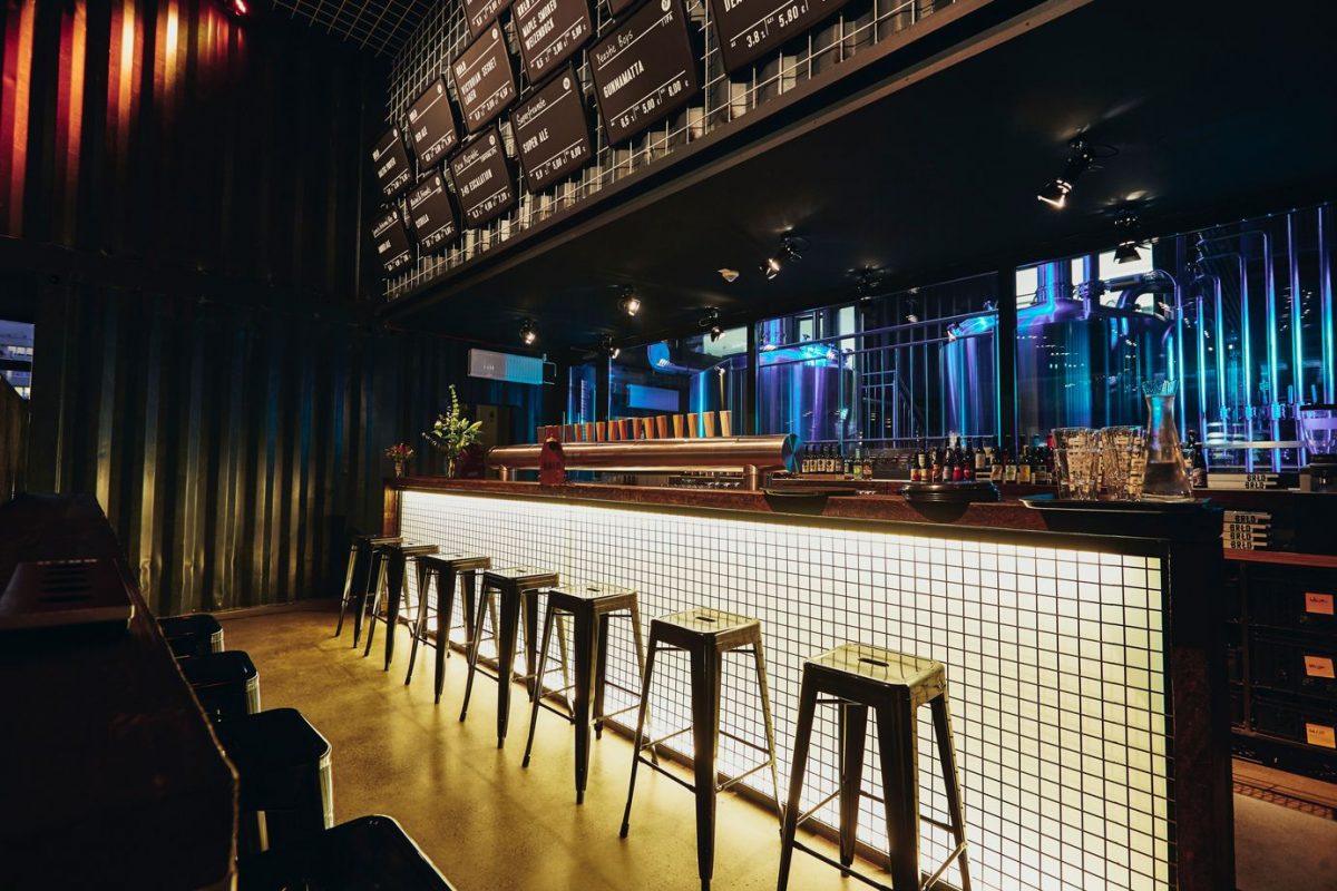 BRLO, Blick auf die Bar im Innenbereich, Barhocker stehen vor einer weißen Kachelwand, bunte Lichter in blau und rot leuchten hinter der Wand