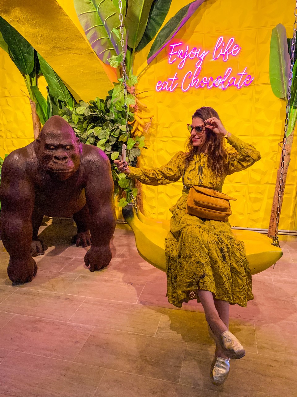 Chocolate Museum Vienna, woman in yellow dress sits on a banana swing. Next to her is a gorilla figure. On the wall is written in illuminated letters 