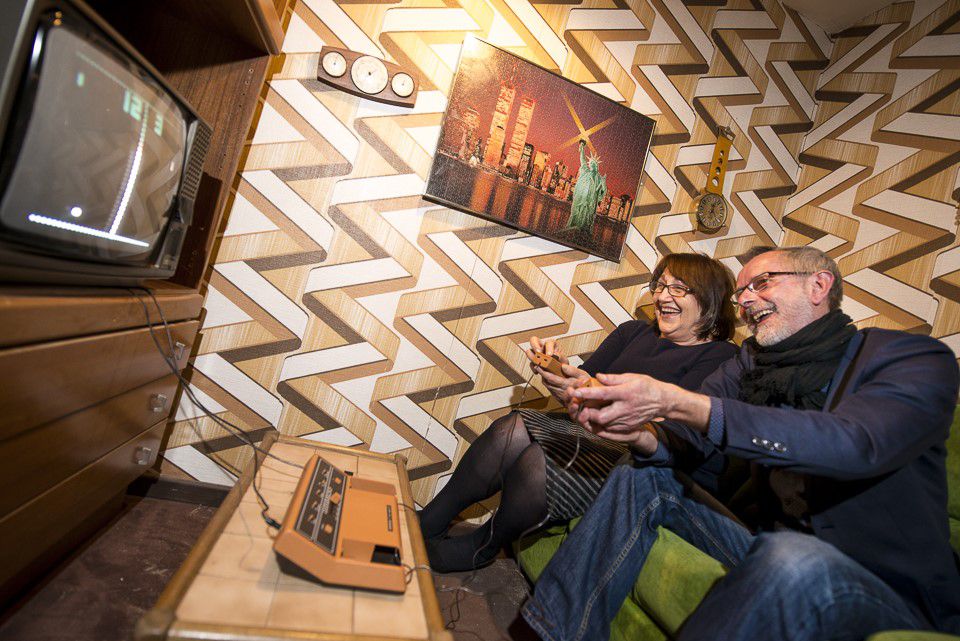 Computer games museum, two people sitting on a green couch playing an old computer game on a TV, environment is designed like an old GDR living room