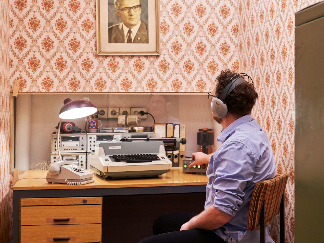 DDR Museum, Abhören, Besucher sitzt am Schreibtisch mit Kopfhörern auf, Bild von Erich Honecker an der Wand