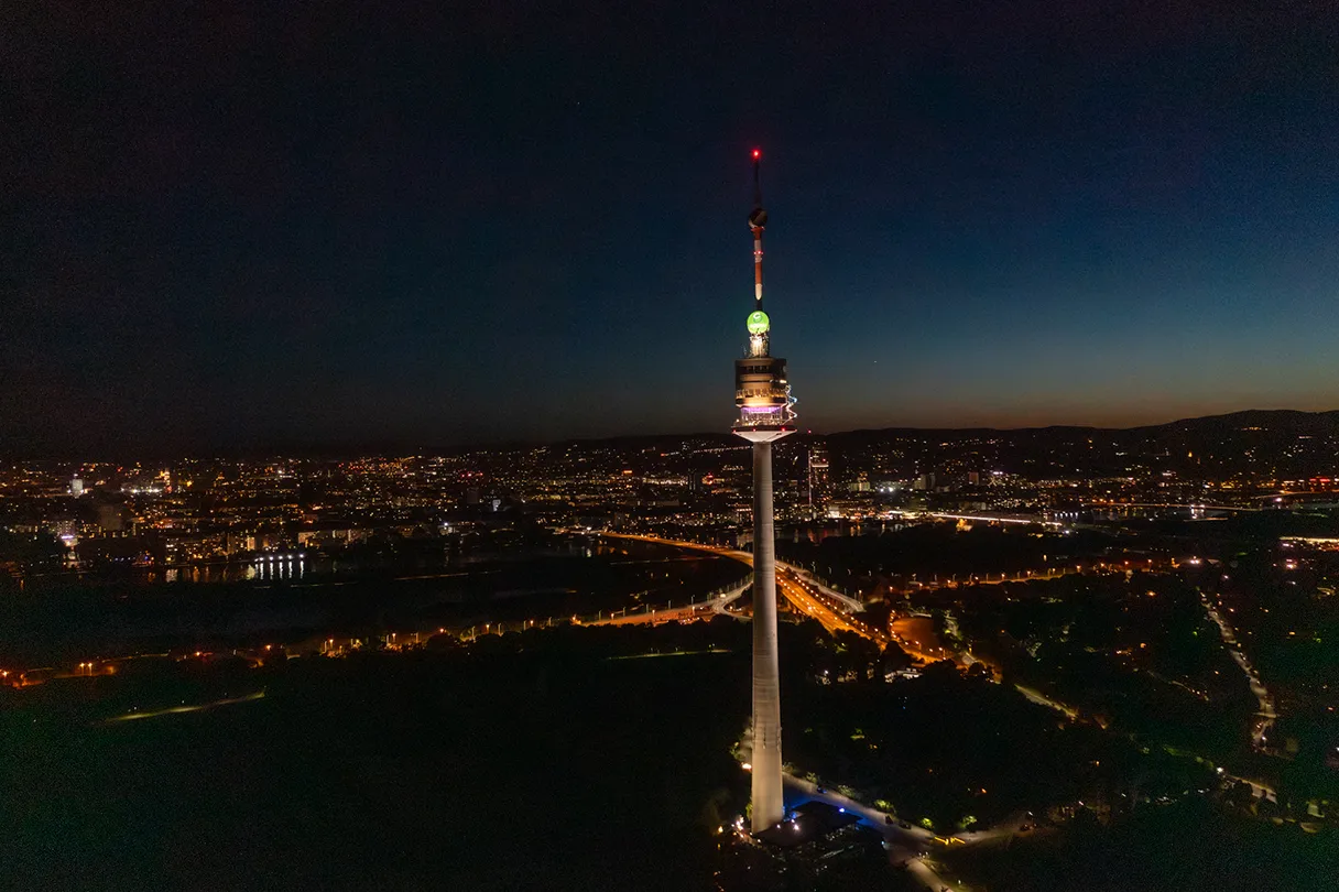 Donauturm. Panoramaansicht auf den Donauturm bei Nacht. Der Donauturm ist angeleuchtet und herum sind die Lichter der Straßen zu sehen.