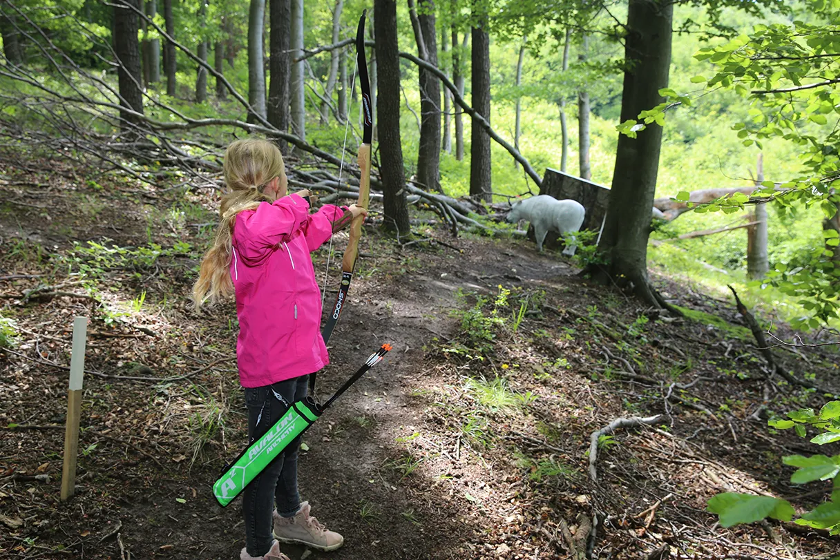 3D Bogensportpark, Erlebniswelt Kahlenberg, Bogenschießen, kleines blondes Mädchen steht im Wald und hat einen Bogen in der Hand, sie zielt auf einen entfernten Hirsch