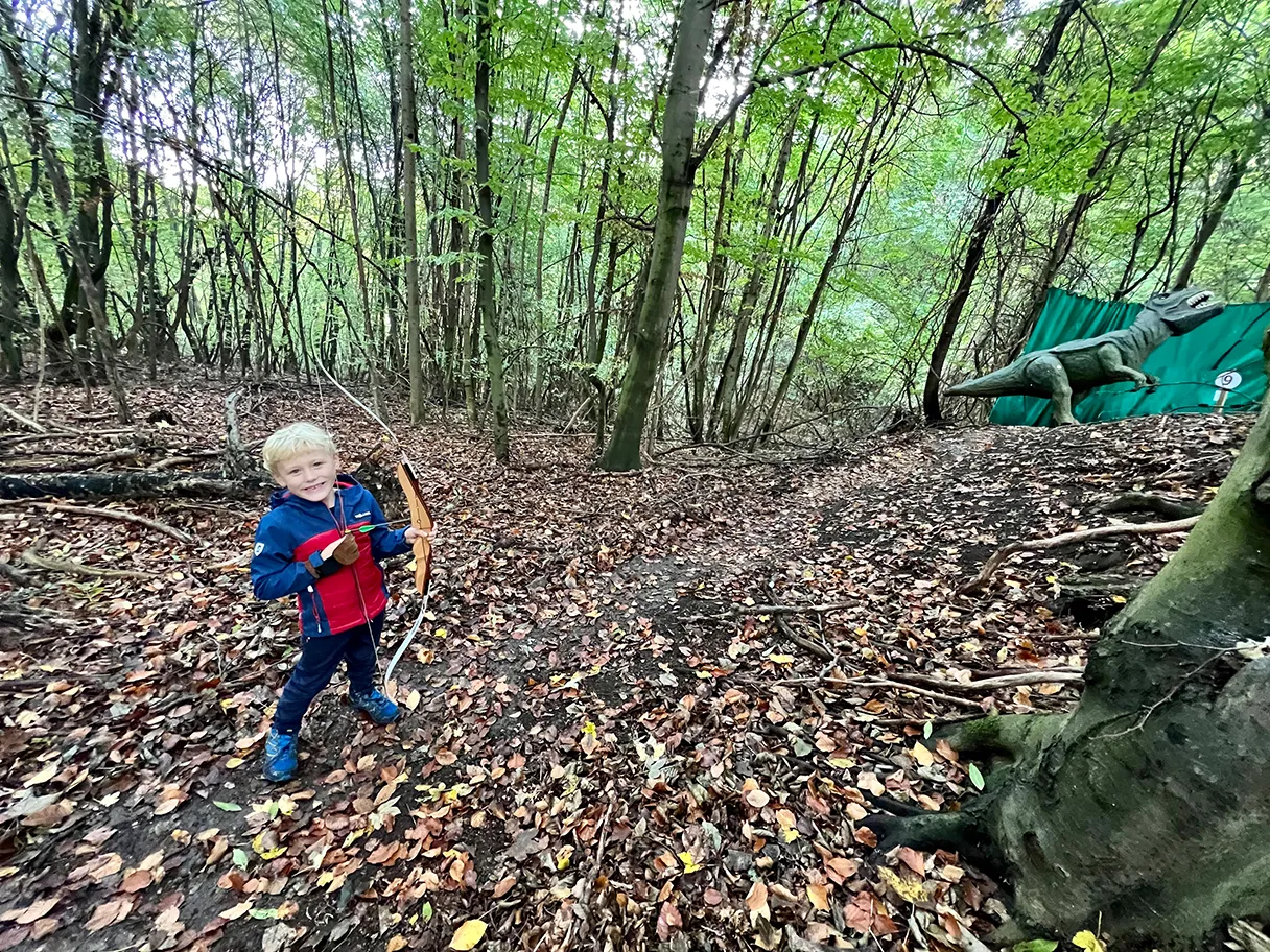 3D Bogensportpark, Erlebniswelt Kahlenberg, Bogenschießen, kleiner blonder Junge steht im Wald und hat einen Bogen in der Hand, im Hintergrund steht ein grüner Dinosaurier