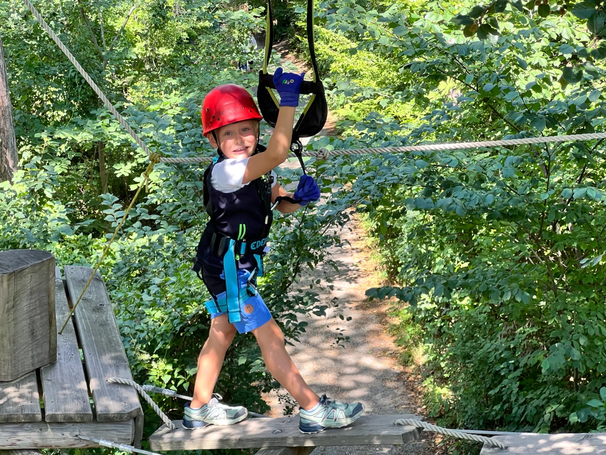 Kleiner Junge hängt an einem Seil, Waldseilpark Parcour, roter Helm auf dem Kopf