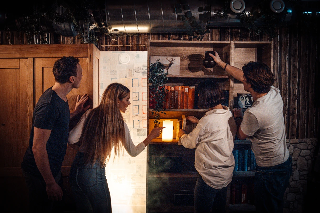 Escape mission, Vienna, four people stand at a bookshelf and try to solve a puzzle, a lamp is burning on the shelf