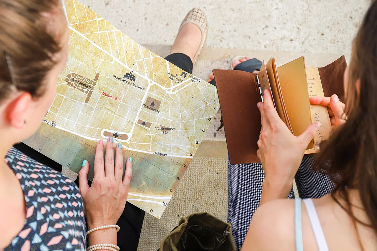 Scavenger hunt, two women sit on the floor and try to solve puzzles, one woman holds a city map in her hand, another holds a notebook, bird's eye view