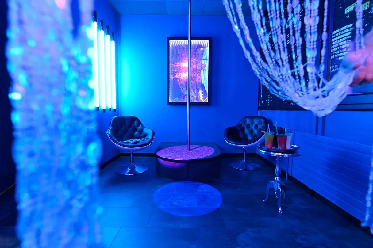 Scavenger Escape Room Vienna, a room illuminated in blue, two armchairs around a table dance pole, curtains and blue neon light