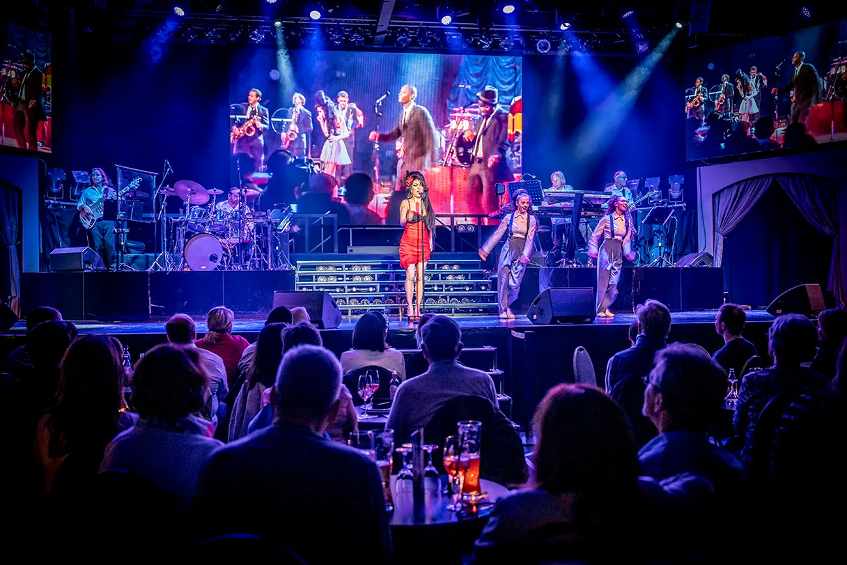 Amy Winehouse Double is on stage and is accompanied by dancers and other musicians on stage. View into the auditorium with a view of the stage. Blue-red stage light.