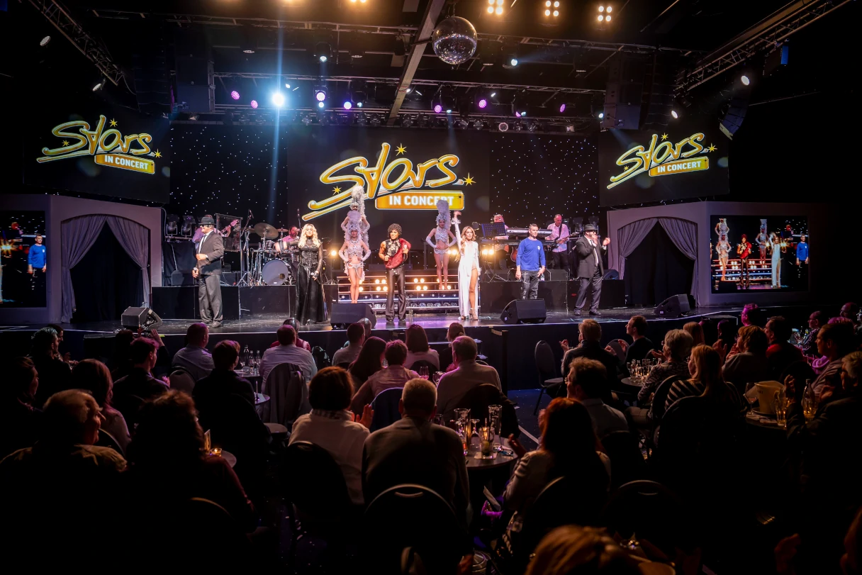 Stars in Concert. View of the show theater. The audience can be seen and various artists are on stage. The yellow Stars in Concert logo can be seen three times in different places on the stage.