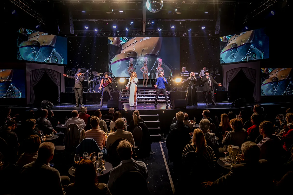 View into the hall and onto the stage. Various artist doubles stand on the stage and perform. Large screens can be seen in the background, providing a graphic backdrop to the show.