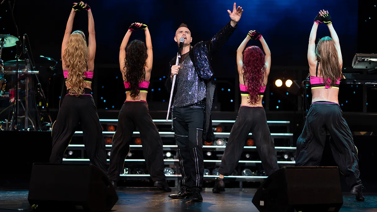 A Robbie Williams lookalike stands on stage and sings with a microphone stand in his hand. Two dancers stand to the right and left with their backs to the audience and stretch their arms in the air.