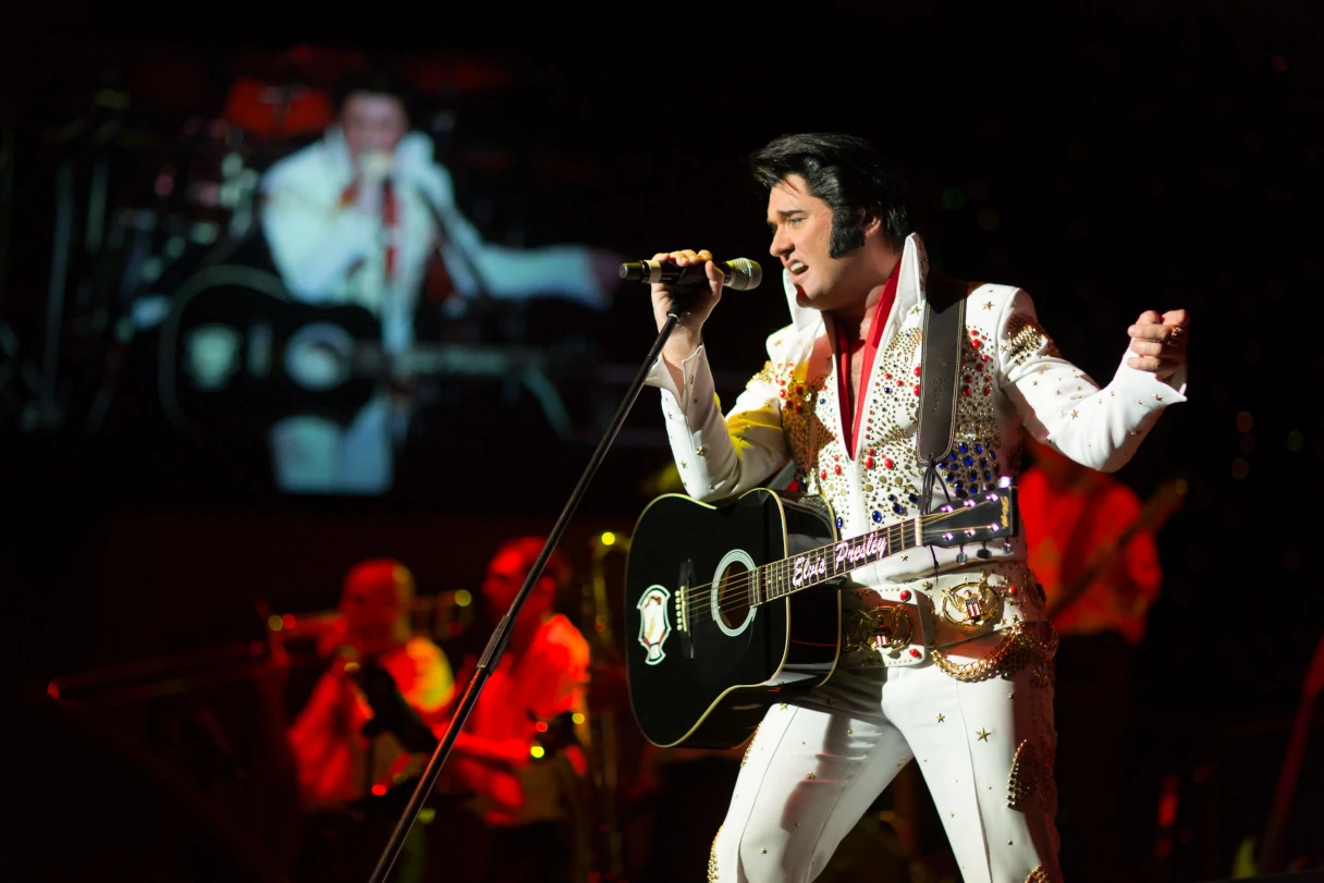 Stars in Concert. An Elvis Presley lookalike stands on stage with a guitar strapped around his neck and sings into a microphone. He is wearing the typical white Elvis suit with the collar turned up.