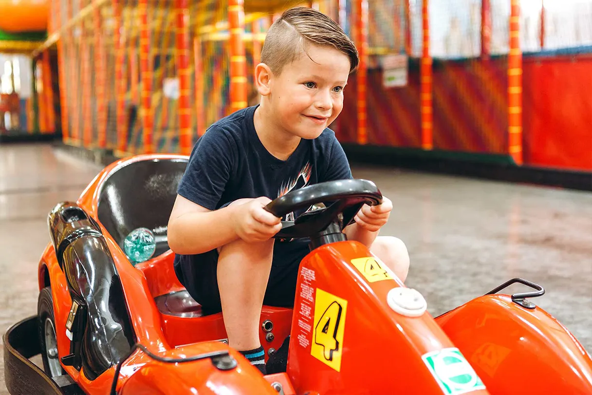 Family Fun, Indoor-Spielplatz, Junge sitzt auf einem orangenen Kart und fährt, er lacht