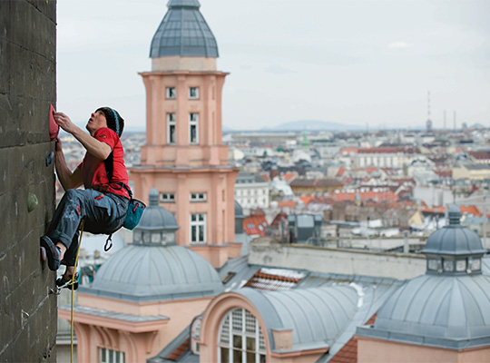 Ein Mann in einem roten T-Shirt hängt an Seilen an der Kletterwand am Flakturm in Wien. Im Hintergrund sind verschiedene Häuser Wiens zu sehen.