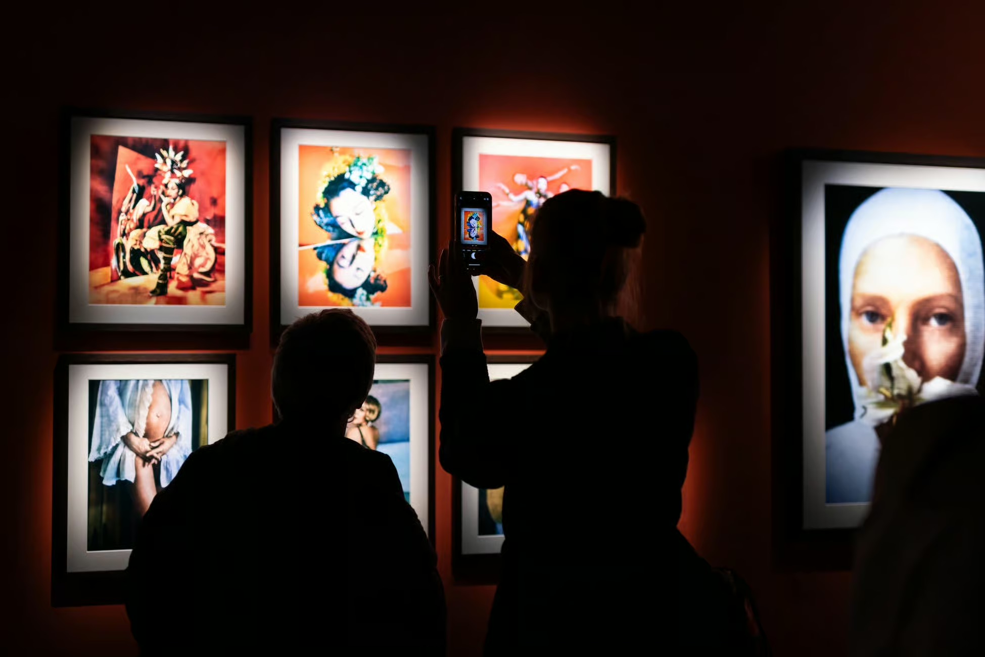 Visitors at Fotografiska Berlin view the exhibition and are visible as black silhouettes. One visitor takes a photo of a colorful picture on the wall with his cell phone.