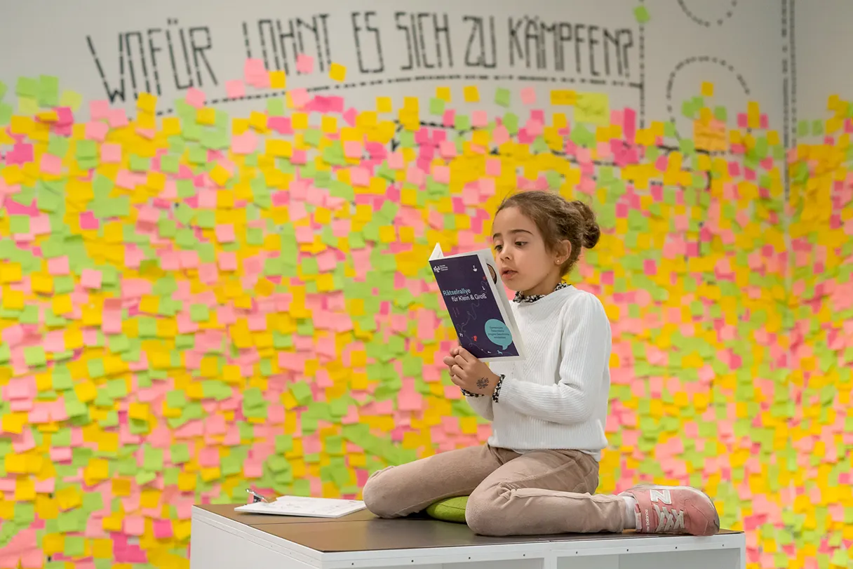 House of History Austria, child sits on a white sitting block and reads in a booklet, the wall behind her covered with colorful Post it notes