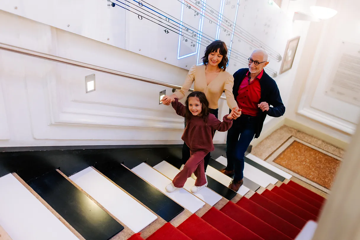 Die klingende Treppe im Haus der Musik Wien. Jede Stufe ist wie eine Klaviertaste und erzeugt einen Ton, sobald man sie betritt. Eine Frau, ein älterer Herr und ein kleines Mädchen laufen zusammen die Treppe hoch und erzeugen verschiedene Klänge.