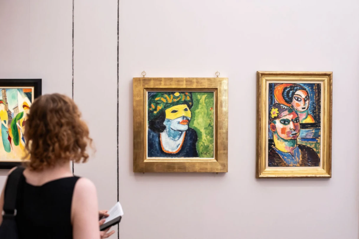 Dauerausstellung KLIMT ⇄ WARHOL. Blick in die Ausstellung. Eine Frau im schwarzen T-Shirt steht mit dem Rücken zum Betrachter und guckt sich Bilder von Pechstein an. Es hängen drei Bilder an der Wand, auf denen bunte Portraits abgebildet sind.