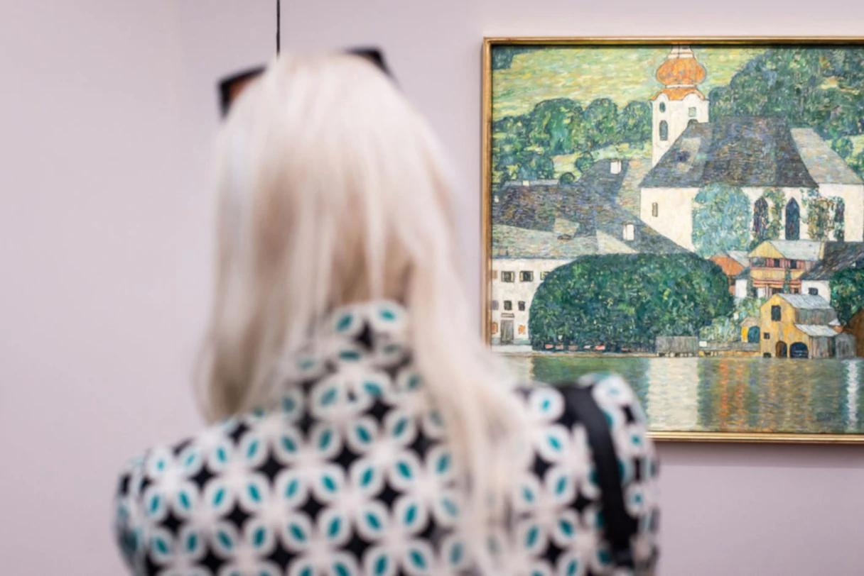 Dauerausstellung KLIMT ⇄ WARHOL. Blick in den Ausstellungsraum. Frau mit langen weißen Haaren, gemusterten Jacke steht mit dem Rücken zum Betrachter und schaut sich ein Bild an.