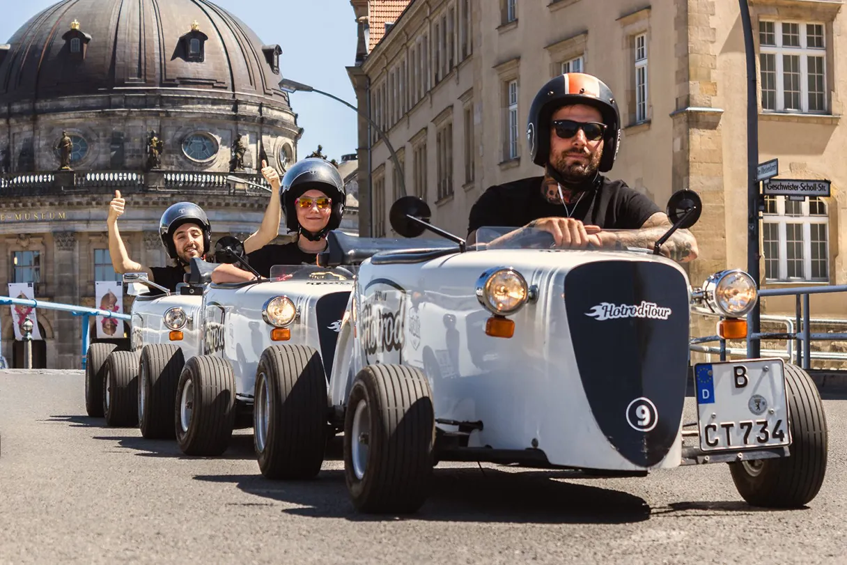 Hotrod tour along the Museum Island. The Bodemuseum can be seen in the background. The hotrods are lined up one behind the other. All drivers are wearing black helmets.