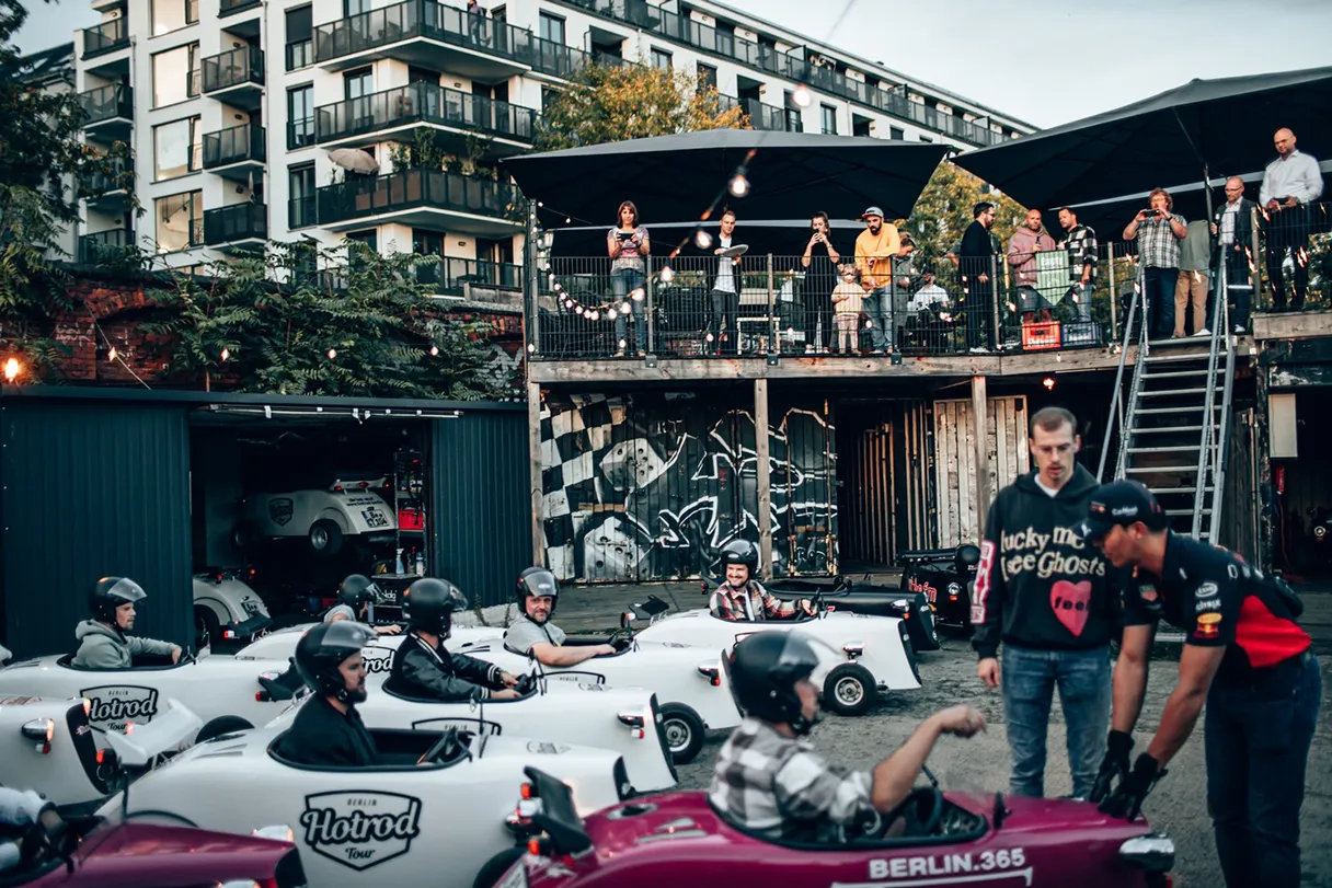 Hotrod Tour group event on the RAW grounds in Berlin. Various white cars are parked next to each other, waiting to get started. There is a terrace on the site where those waiting can enjoy pizza and drinks while they wait.