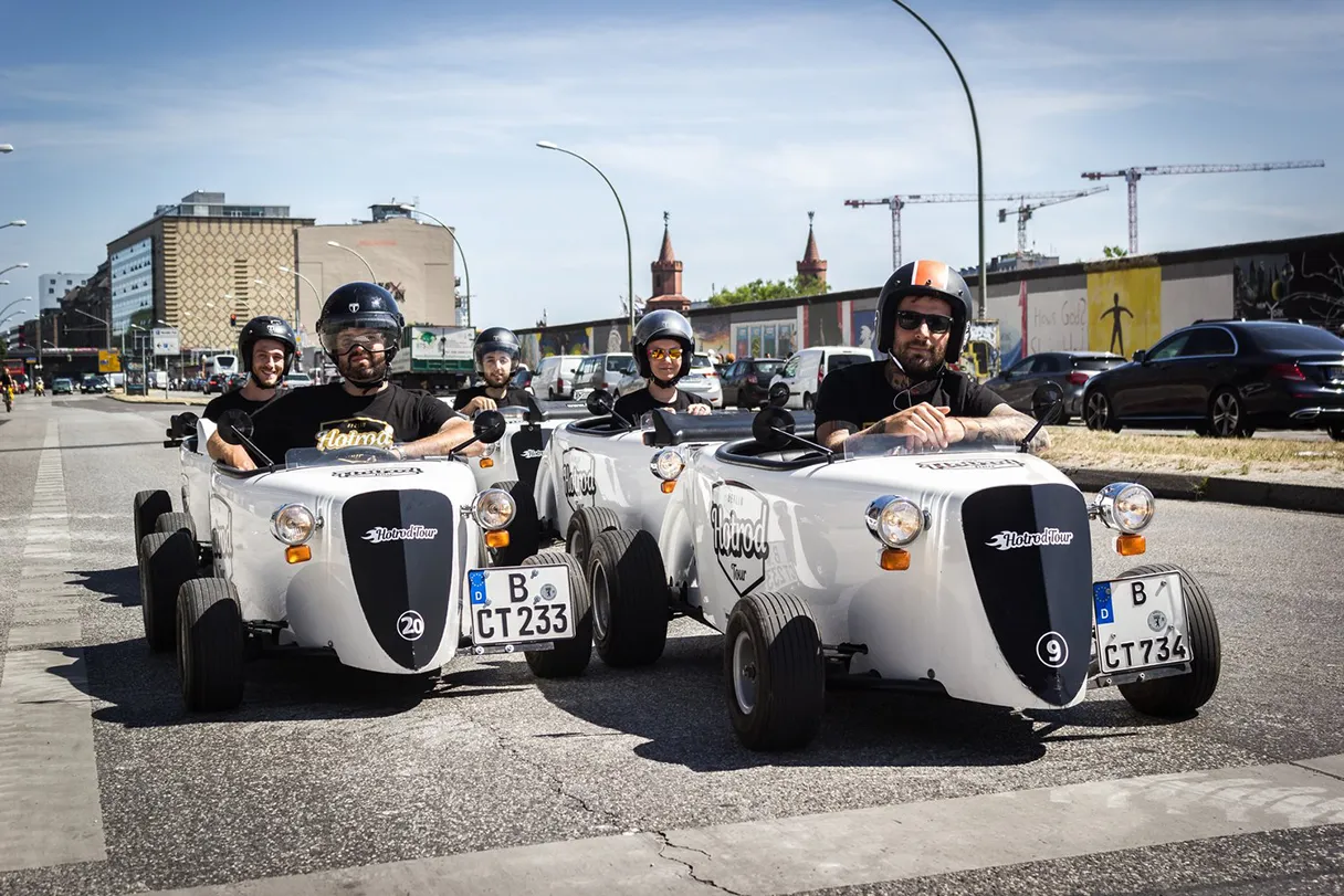 Hotrod tour at the EastSide Gallery. Five hotrods drive on the street and look into the camera.