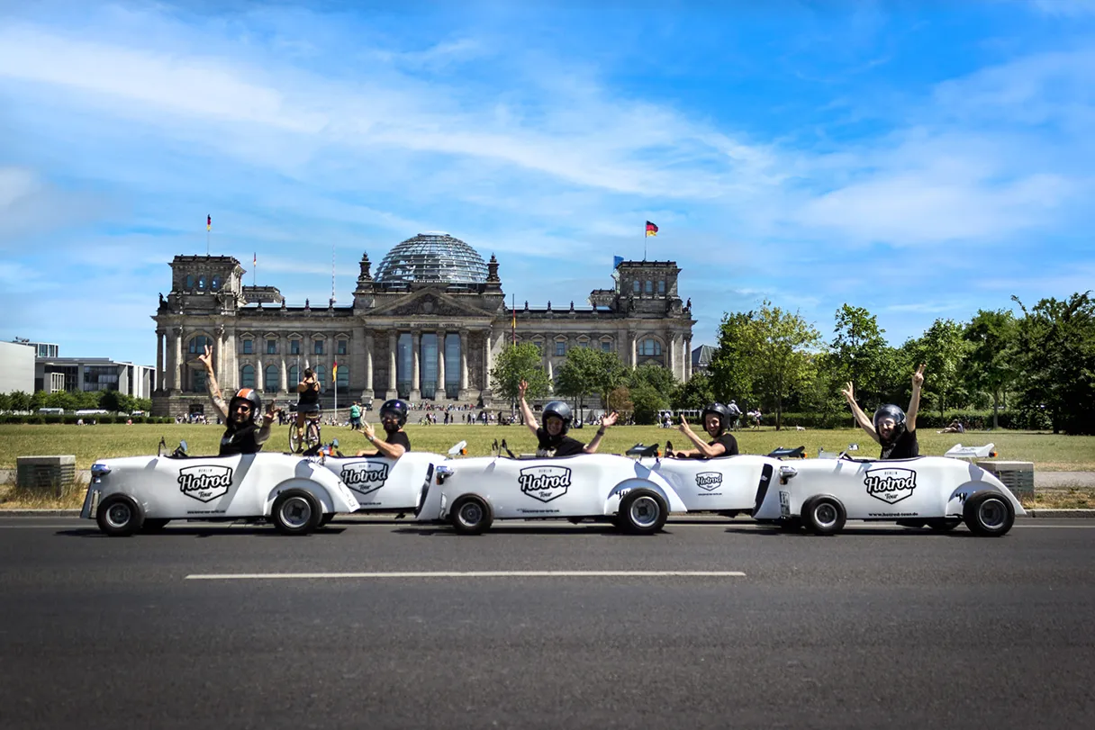 Hotrods are lined up in front of the Reichstag. Blue sky, a few hazy clouds, green lawn.