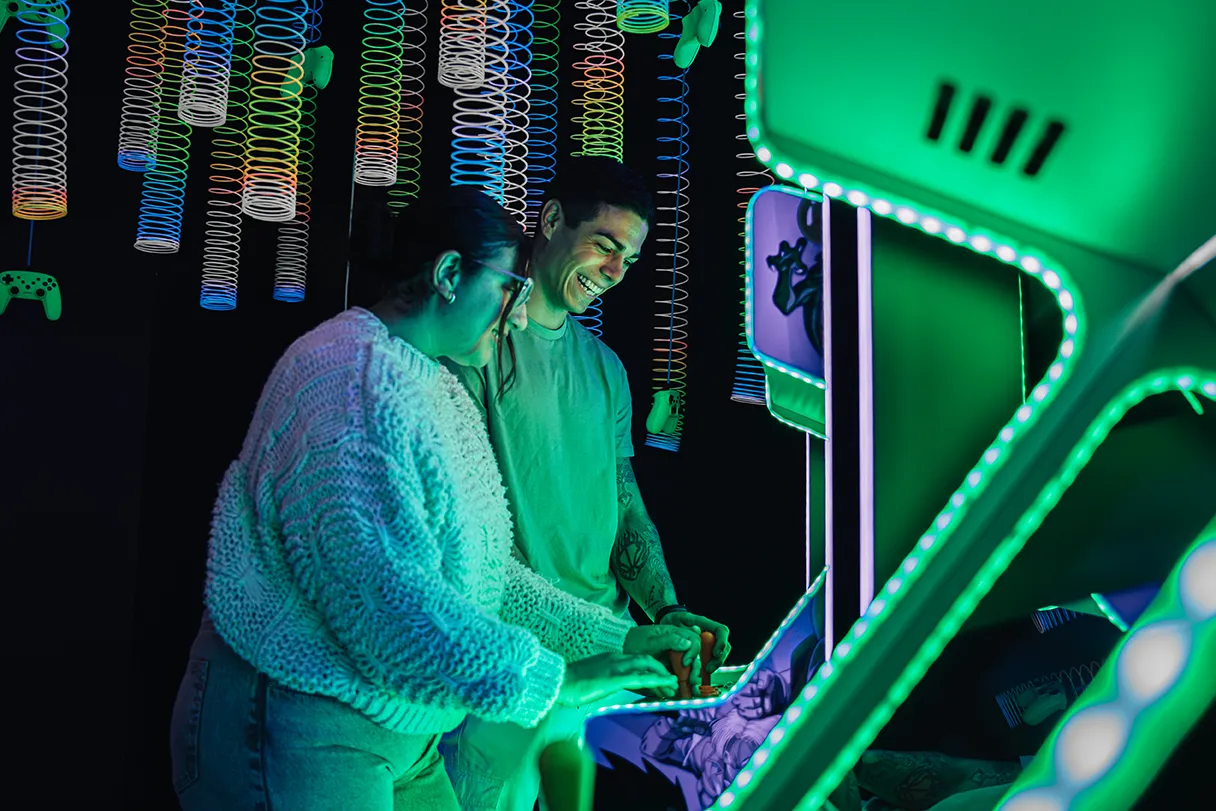 IKONO Berlin, two visitors stand at arcade slot machines, colorful neon spirals light up around them