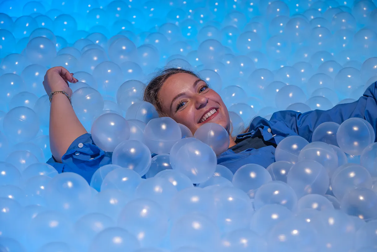 IKONO Berlin, woman lying in a ball pool with transparent balls, only her head and arms are visible, the rest is covered by balls