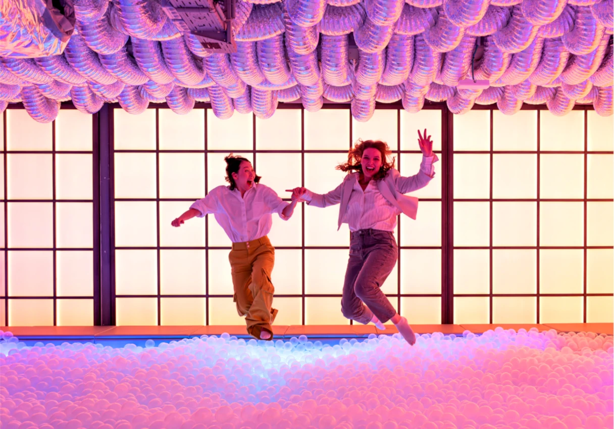 IKONO Vienna, two women jump hand in hand into a large ball pool with white balls