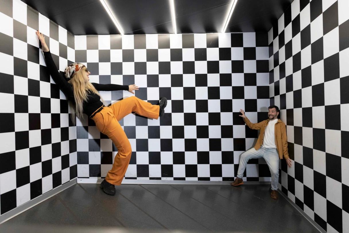 Illuseum Berlin, Ames Room, woman stands against a black and white checkered wall, in another corner sits a man who looks much smaller