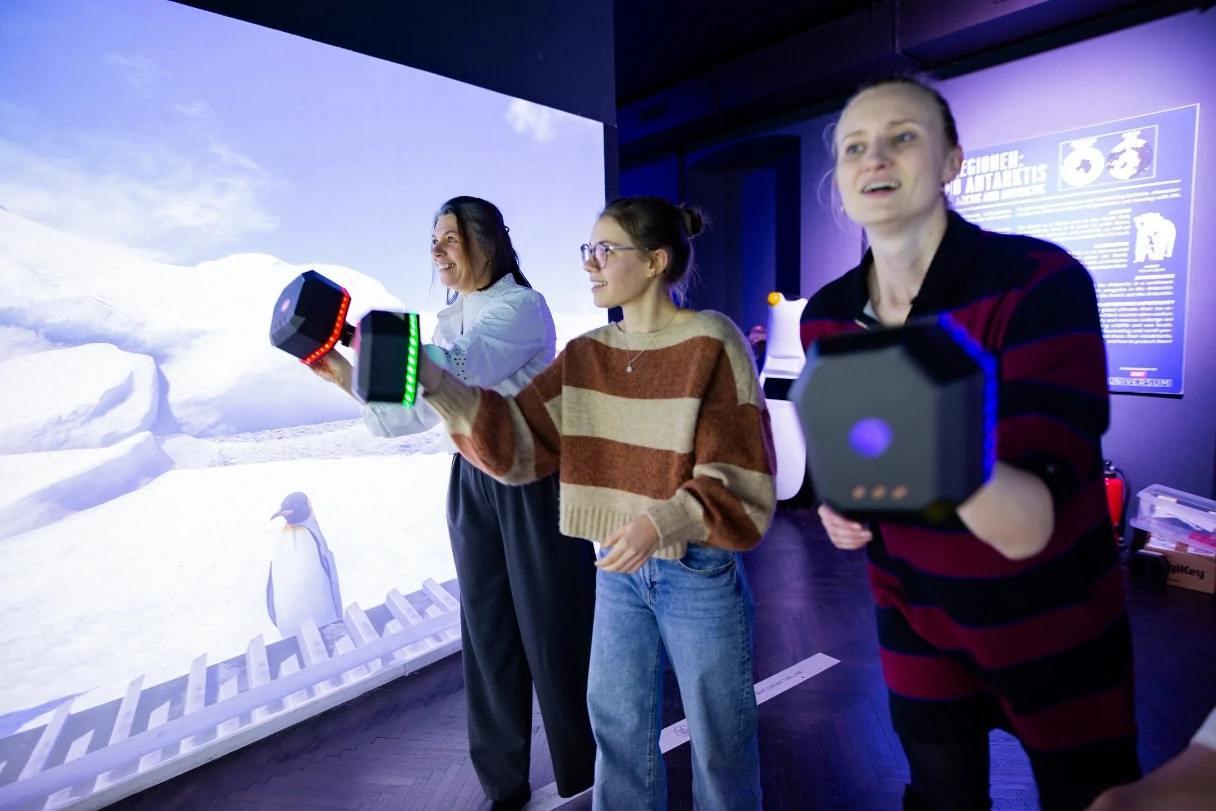 Drei Besucherinnen des Immersium:Wien haben achtkantige Geräte in den Händen, mit denen man Dinge auf der großen Leinwand bewegen kann. Es ist wie ein großes Computerspiel. Alle drei Frauen lachen. Im Hintergrund ist ein Pinguin auf der Leinwand zu erkennen.