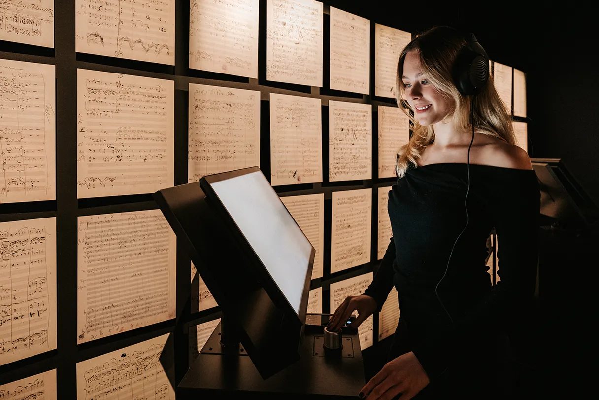 Johann Strauss Museum, view of the exhibition. A visitor stands at a terminal and composes her own waltz. She is smiling. Various scores hang on the walls.