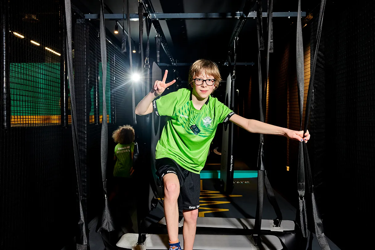 Jumpworld One, boy with glasses in green T-shirt and black pants shows the peace sign with his fingers, standing on a trampoline