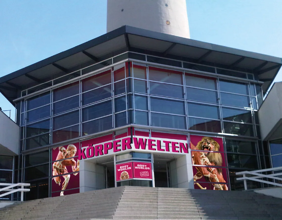 Außenansicht des Eingangsbereiches des KÖRPERWELTEN Museum Berlin. Das pinke Logo und zwei Grafiker von Exponaten sind rechts und links der Türen zu erkennen. Treppen führen zum Eingang.