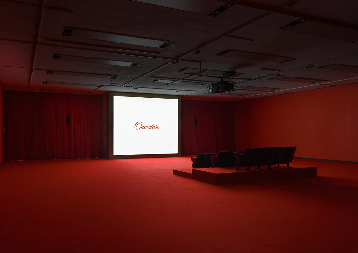 View of the exhibition space. Red carpet, red curtain, a few seats on the right-hand side. It looks like a movie theater. A bright screen with something written on it in red letters. There are no people in the room.