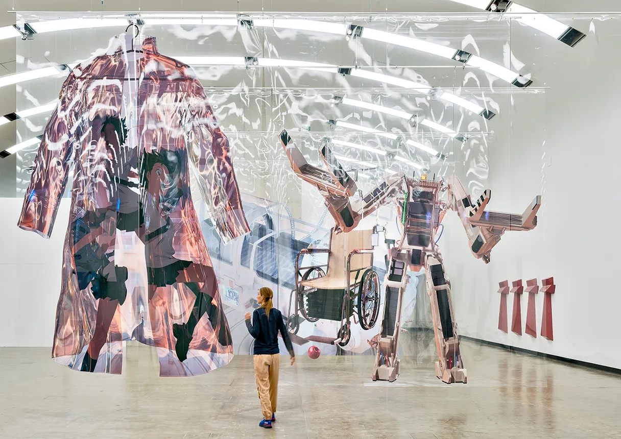 View of the exhibition. Various works of art hang from the ceiling. A coat can be seen on the left. A woman in a black sweater and yellow trousers stands in front of it and looks at the artwork.