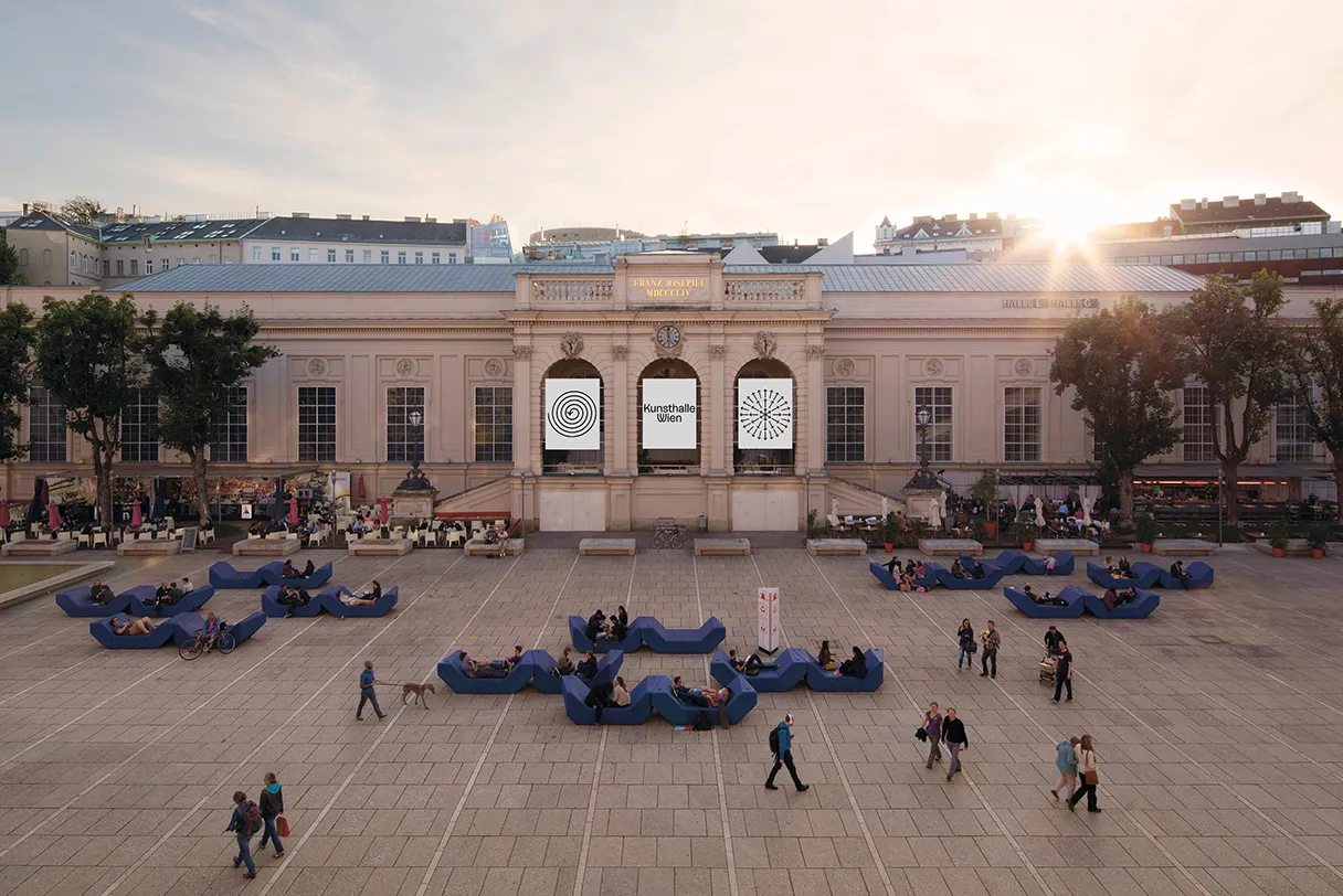 View of the MuseumsQuartier Vienna from above. The sun is slowly setting on the right-hand edge of the picture, people are walking across the square and there are dark purple sofas to relax on.