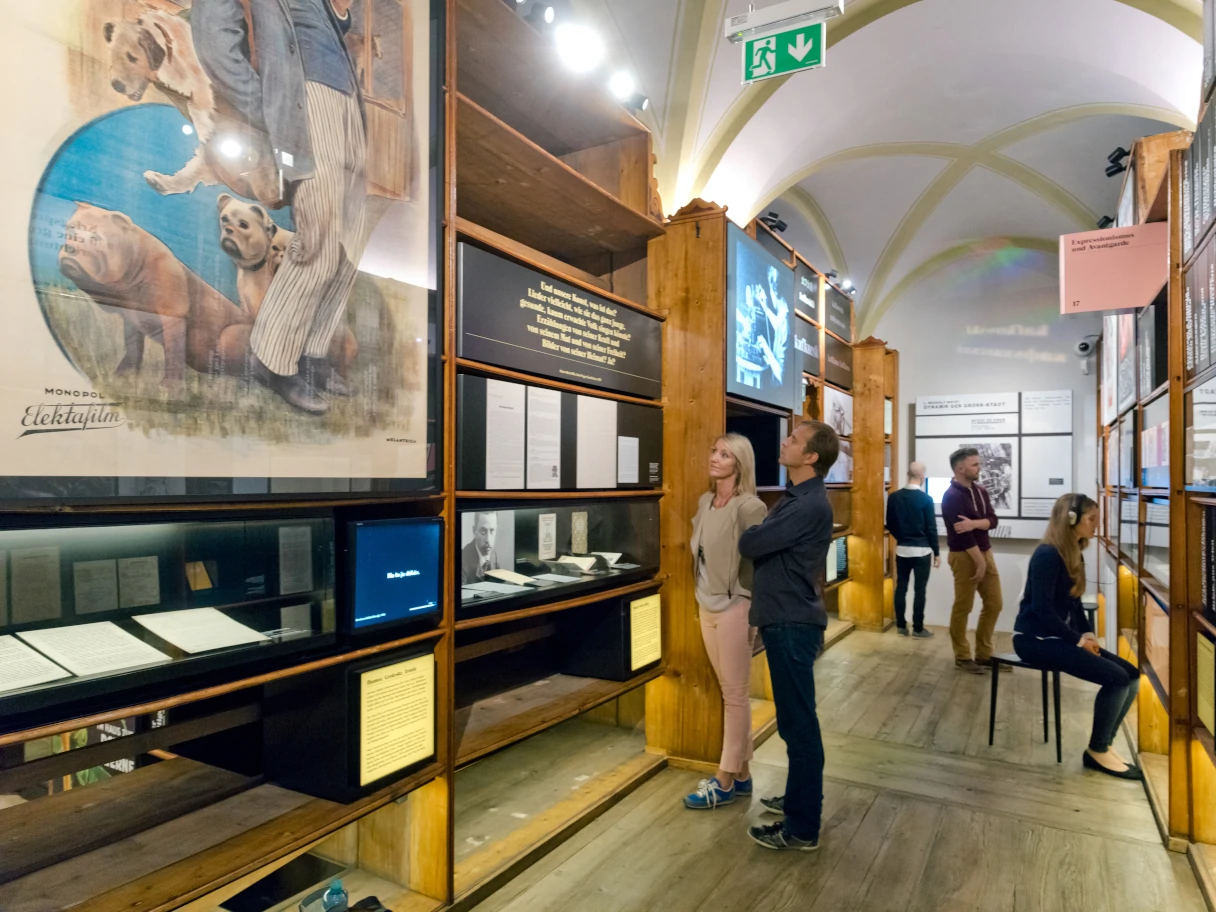 Literaturmuseum Wien. Blick in die Ausstellung. Besucher stehen vor unterscheidlichen Ausstellungsstücken und lesen sich die Erklärungen durch. Verschiedene Regale rechts und links im Bild.